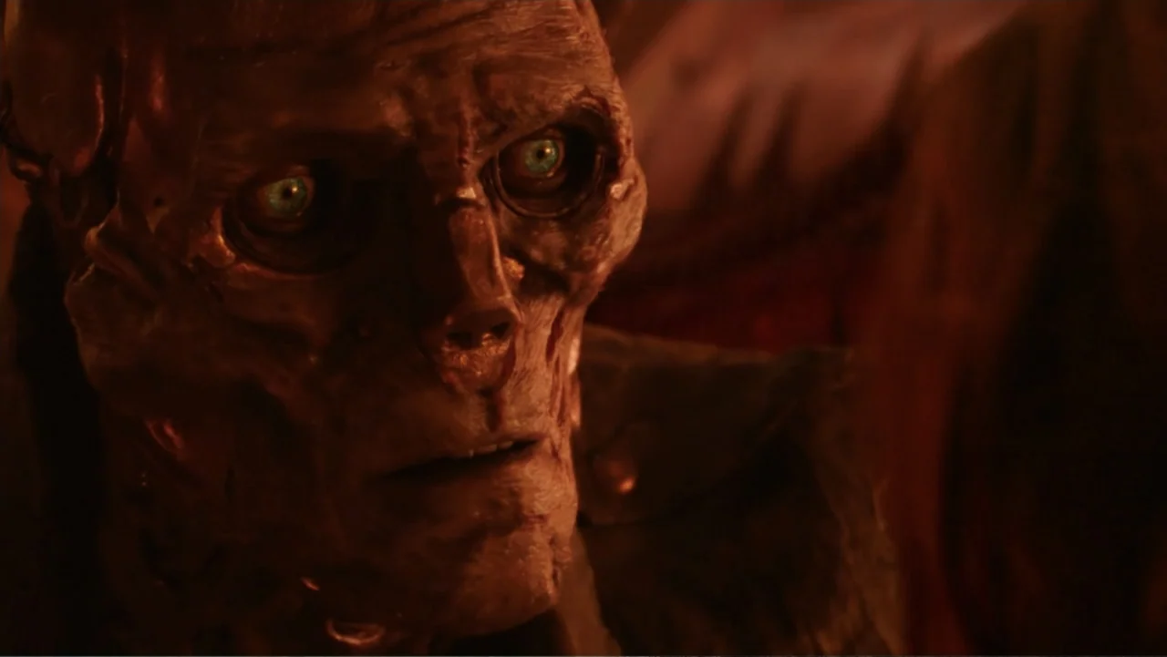 Mortal Engines (2018) movie still: closeup — A dark interior with warm firelight casting shadows on a synthetic humanoid face. The subject gazes…