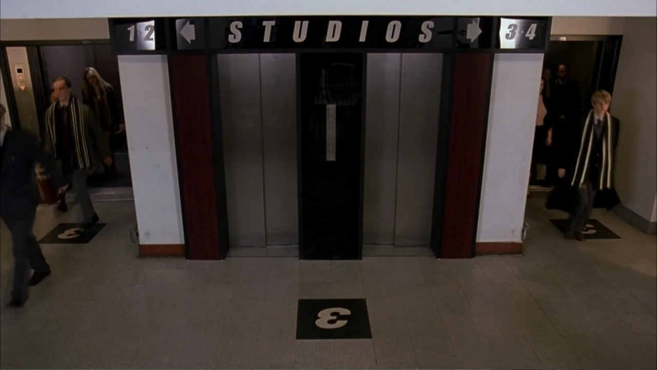 Starter for 10 (2006) movie still: wide — A sterile building lobby features two sets of elevator doors with studio direction signs. Several people…