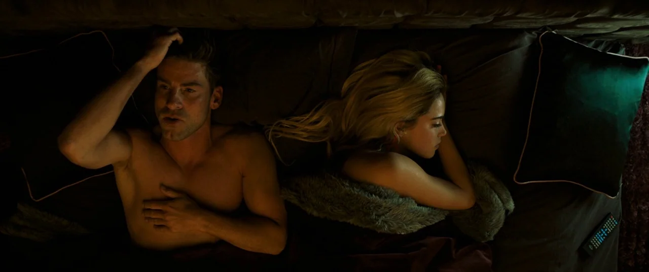 Revenge (2017) movie still: medium — A man and a woman lie in a dark, plush-lined bed at night. The man remains awake while the woman sleeps, with a…