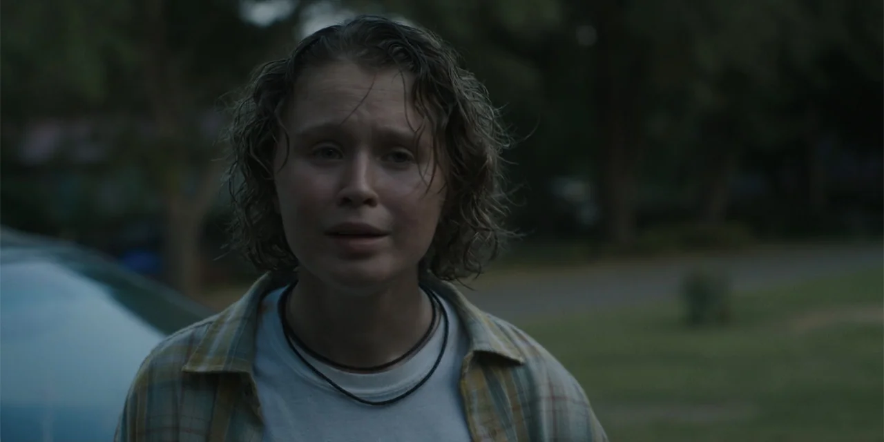 Caddo Lake (2024) movie still: medium — A residential street at dusk with blurred greenery and trees in the background. A woman with wet hair stands…