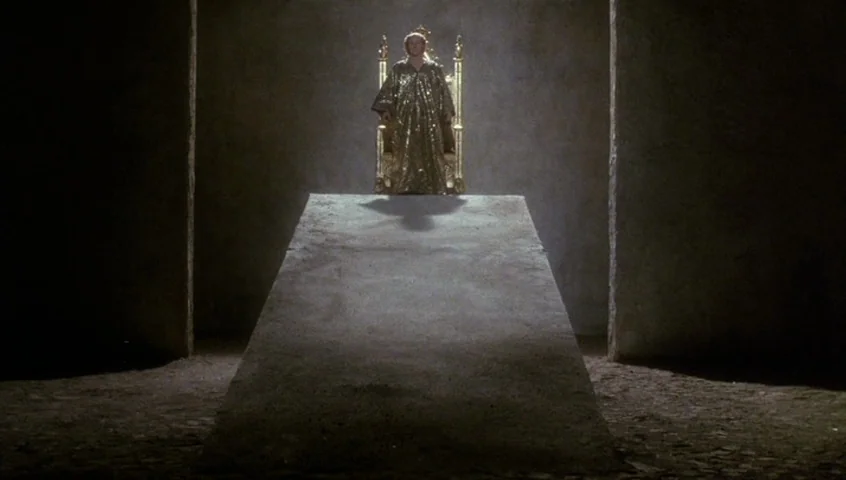 Edward II (1991) movie still: wide — A dark stone room contains a central raised platform shaped like a wide stone ramp. A figure in a gold robe…