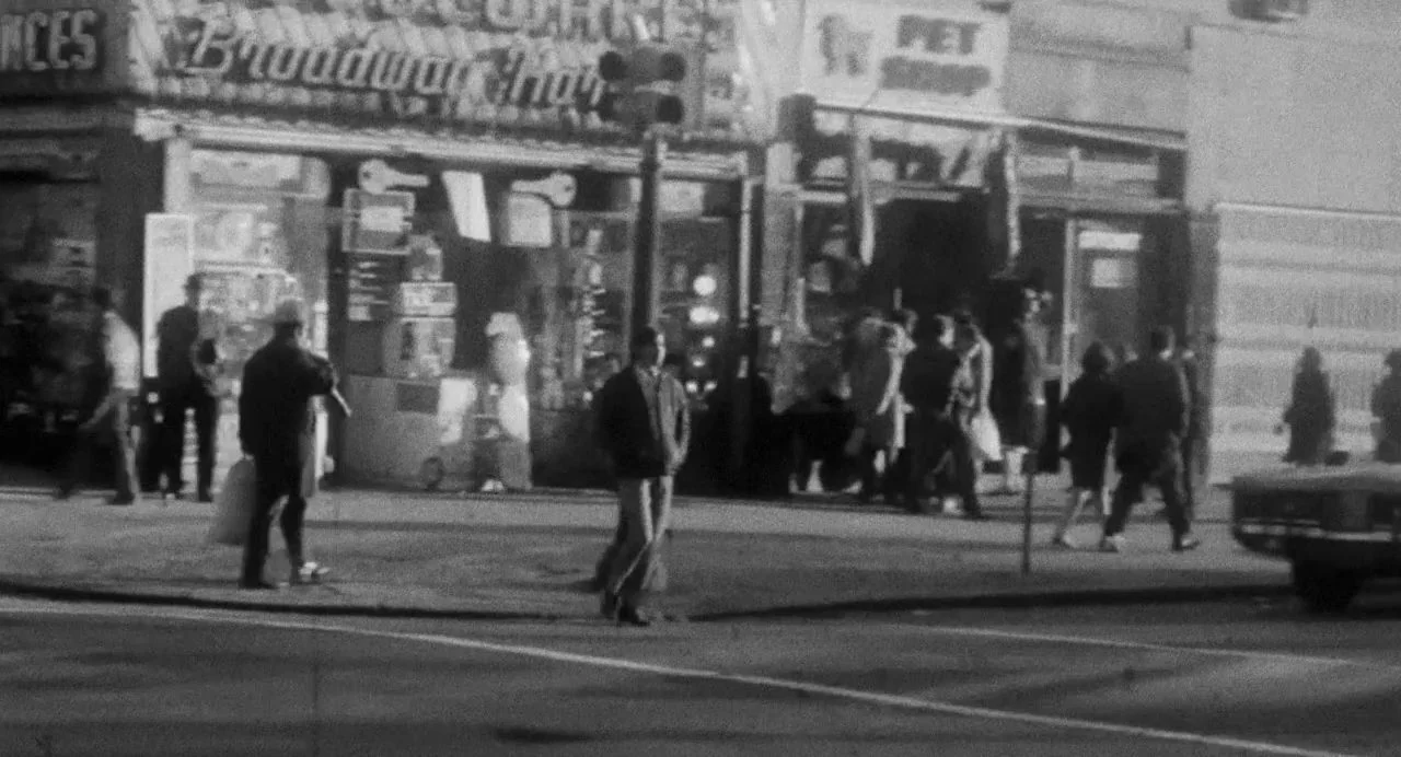 Song of Granite (2017) movie still: wide — A city street at night features several storefronts with visible signs for shops. People walk on the…