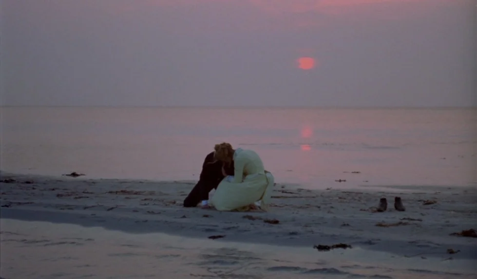 Elvira Madigan (1967) movie still: wide — A quiet sandbar extends into a calm sea under a muted sunset sky. Two figures are huddled together on the…