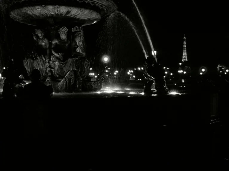 Eloge de L’amour (2001) movie still: wide — A dark stone fountain with figurative carvings sits in a public square at night. The Eiffel Tower is…
