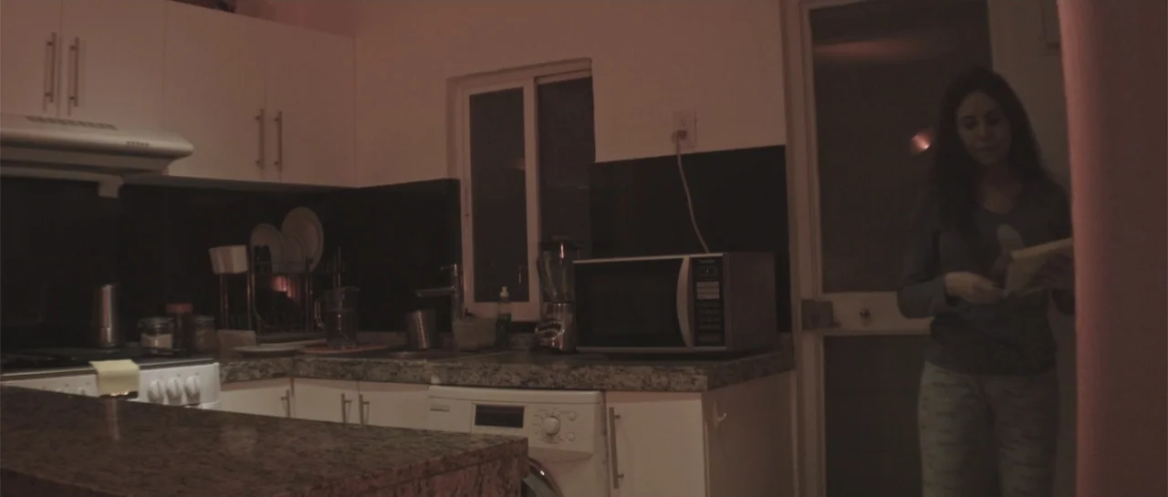 Amor Mutante (2019) movie still: medium — A dimly lit contemporary room contains white cabinets and a granite countertop. A young woman stands in…