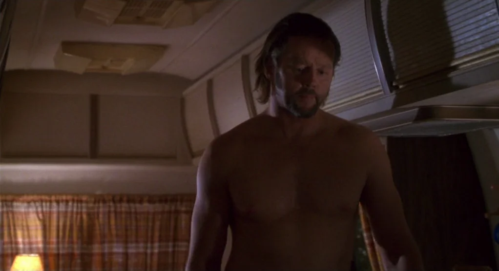 The Crossing Guard (1995) movie still: medium — The interior of an RV has tan cabinets and a plaid curtain over the window. A shirtless man with a…