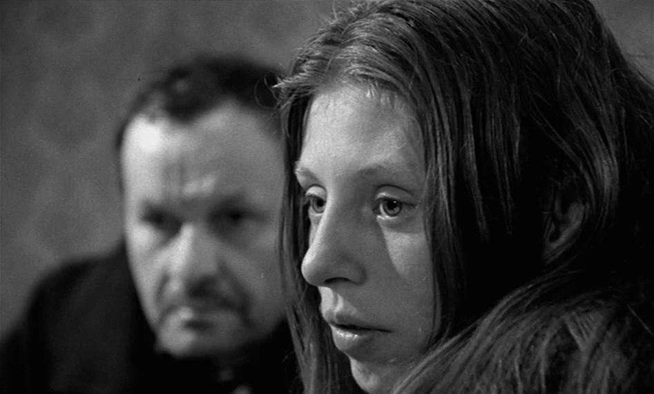 The Man From London (2007) movie still: closeup — The frame shows a young woman in the foreground and a man behind her. They occupy a dark indoor…