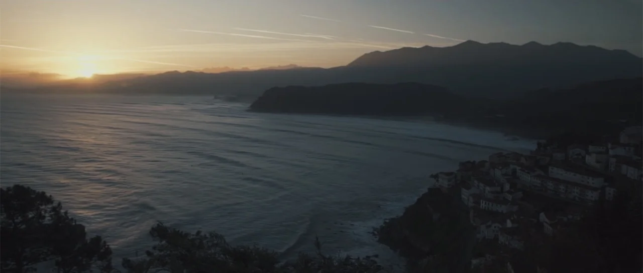Taking Back the Waves (2017) movie still: establishing — A coastal village rests on a cliffside overlooking a large body of water. The sun sets…