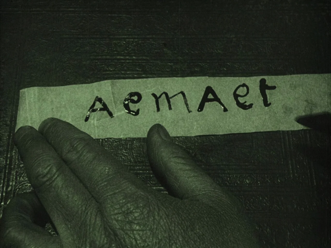 The Golem (1920) movie still: closeup — A close view shows a hand resting on a dark, textured, embossed surface. A slip of paper with the…