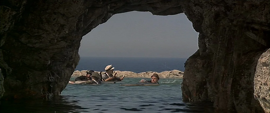 Popeye (1980) movie still: wide — A view from inside a dark limestone sea cave looking out toward the ocean. Four men in period hats float in the…