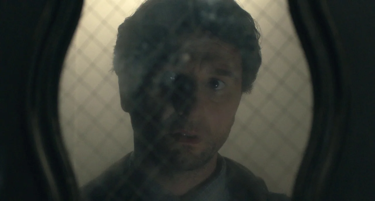 A Beautiful Day In The Neighborhood movie still: closeup — A man peers through a wired glass window set within a dark frame. He stares outward from…