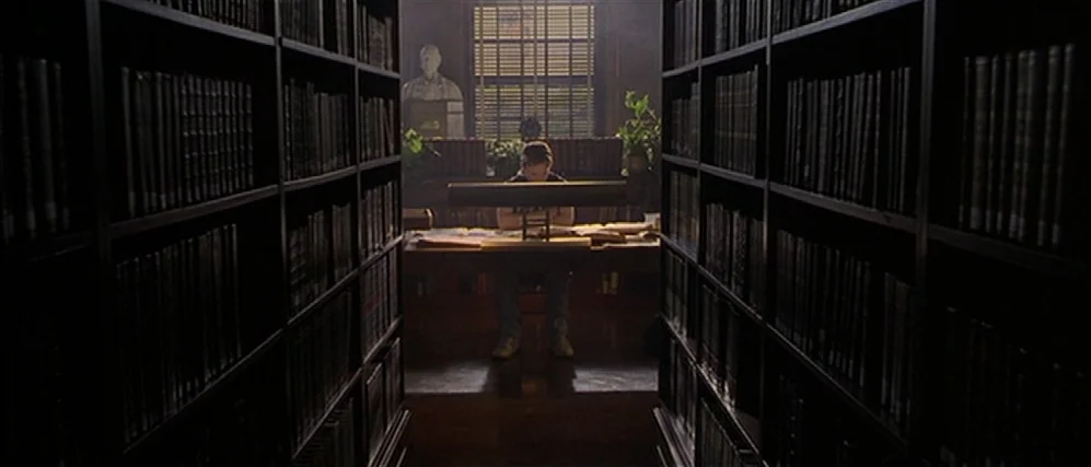 Apt Pupil (1998) movie still: wide — A dark, symmetrical hallway of tall bookshelves leads toward a study area at the back. A person sits at a desk…