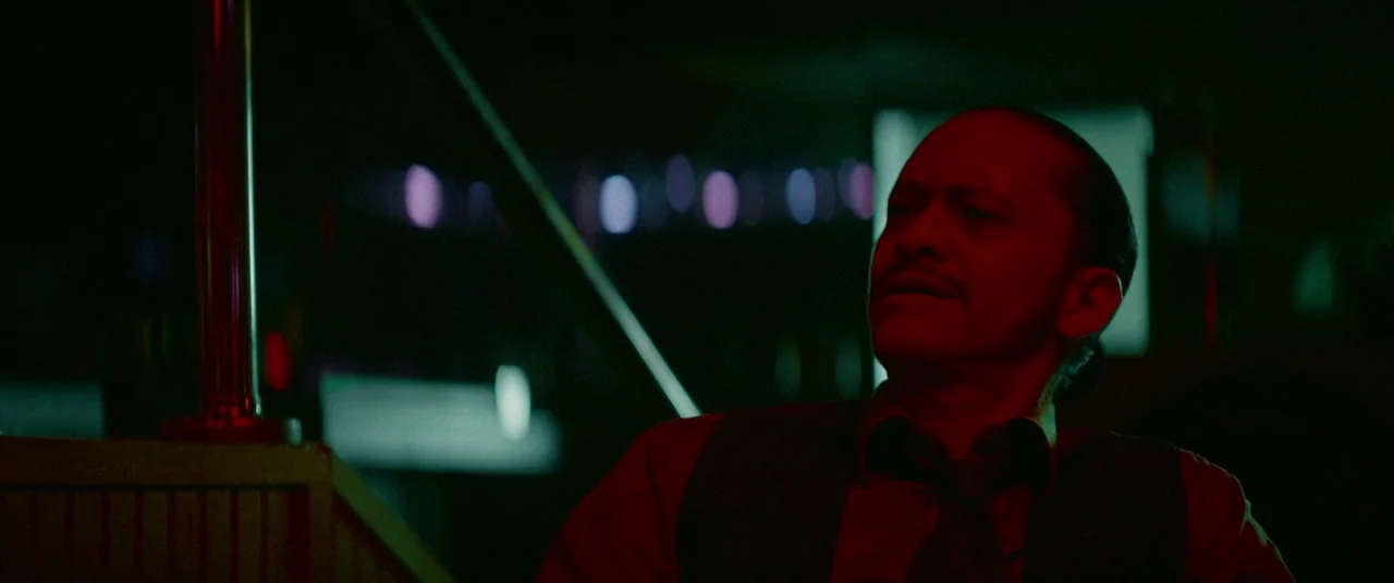 Triple 9 (2016) movie still: medium — A dark venue is illuminated by red and green ambient neon light. A man wearing a vest and tie sits while…