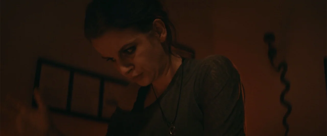 Mennonite's Bite (2023) movie still: medium — A dimly lit interior space is obscured by heavy shadows and a warm color cast. A young woman with dark…