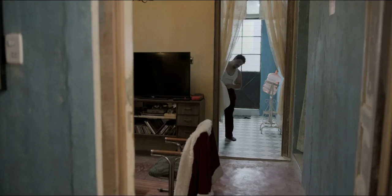 El sabor de la Navidad (2023) movie still: medium — An interior living space features a television, a desk, and a doorway beyond. A man stands in…