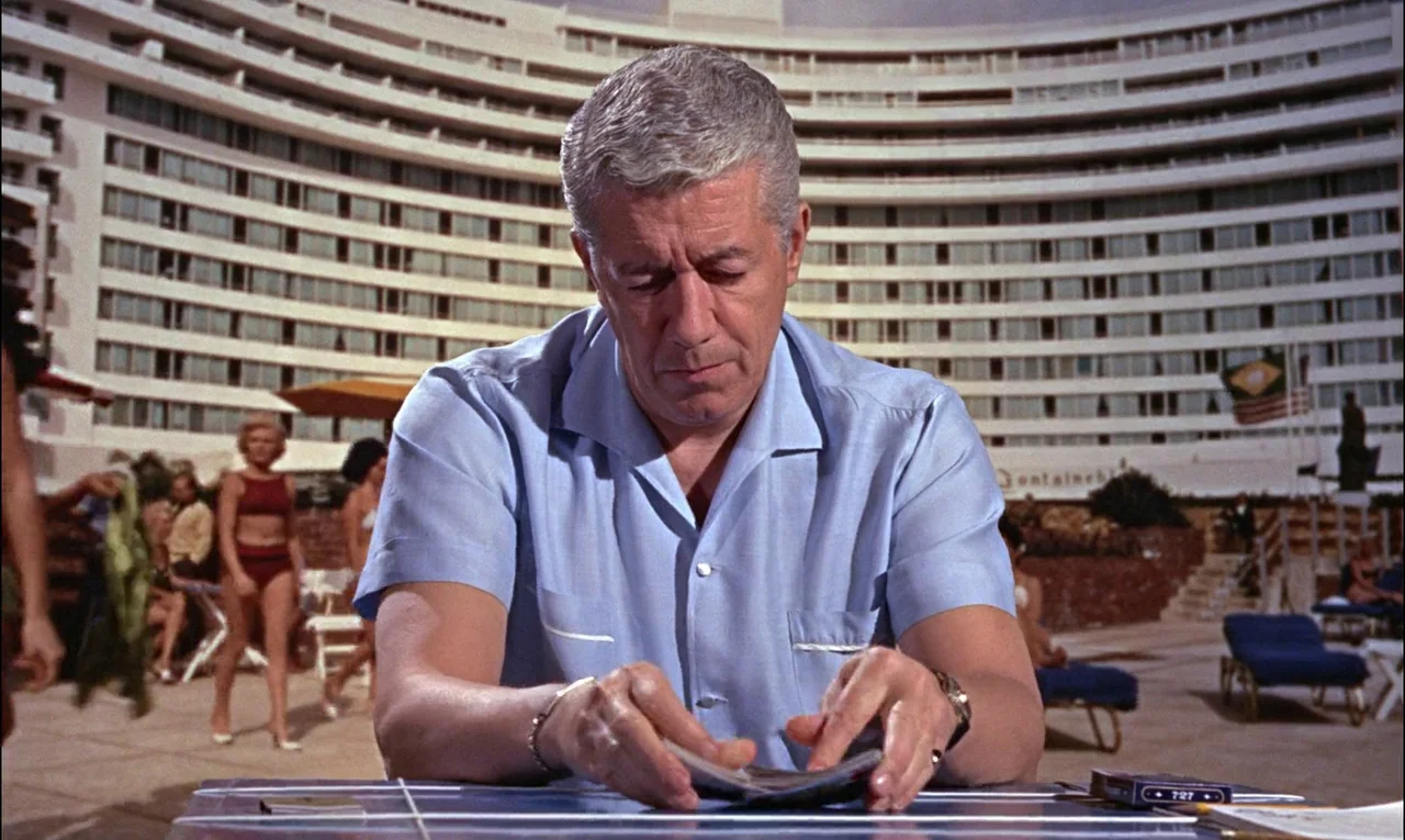 Goldfinger (1964) movie still: medium — A man sits at a tiled table with a large, curved hotel building background. He is shuffling a deck of cards…