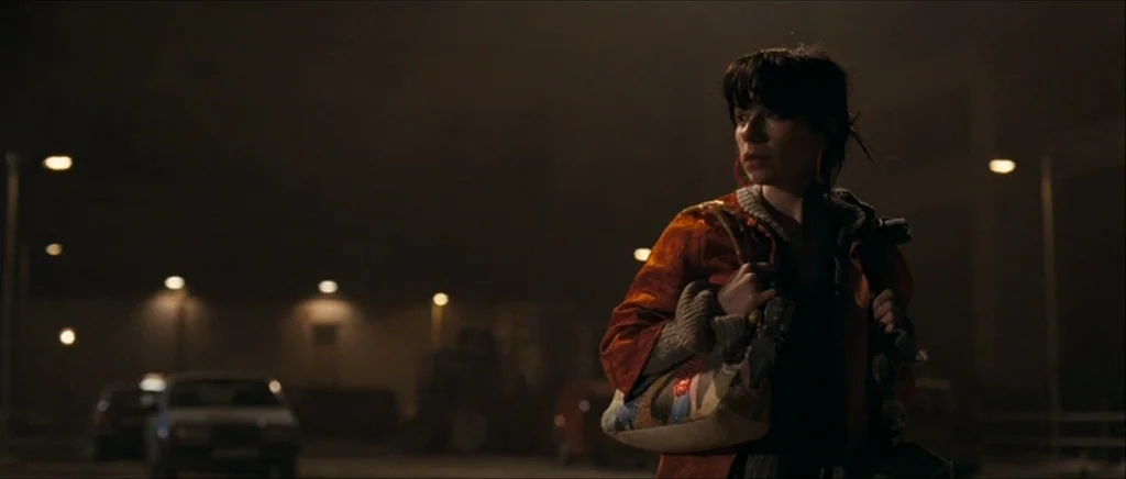 Happy-Go-Lucky (2008) movie still: medium — A dark, dimly lit parking lot is illuminated by sparse, warm street lighting at night. A woman in an…