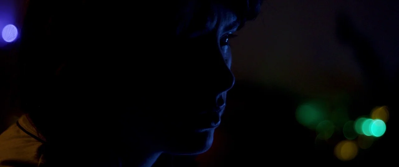 The Canal (2014) movie still: closeup — A dark interior space at night with colored light bokeh visible in the background. A woman's face is…