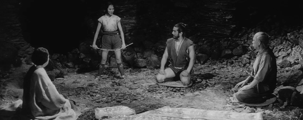 The Hidden Fortress (1958) movie still: wide — A dark stone cave with rough walls and a rocky, uneven ground surface. Four individuals are…