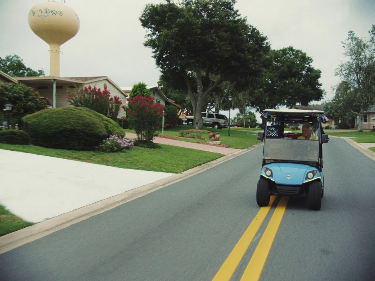 Some Kind Of Heaven (2020) movie still: wide — A suburban street in Florida features well-manicured lawns, a large water tower, and residential…
