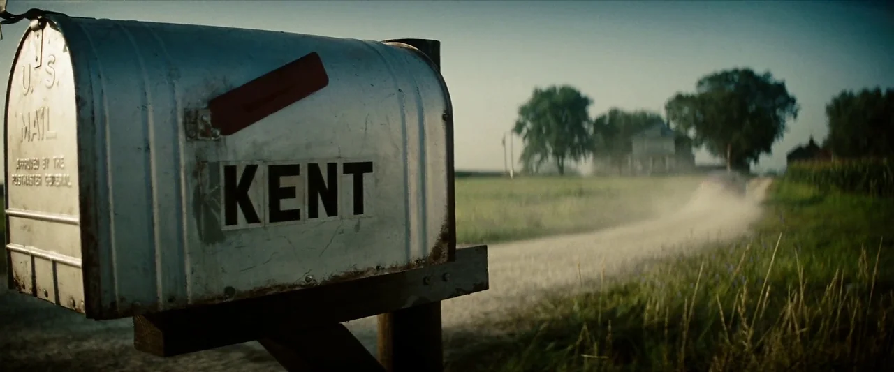 Man Of Steel (2013) movie still: closeup — A metal mailbox with the name KENT is in the immediate foreground. In the distance a car travels along a…