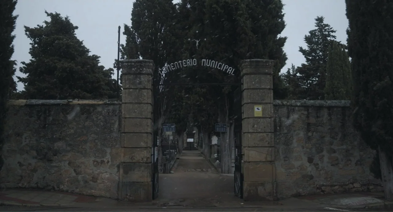 Los días azules (2025) movie still: wide — A stone masonry gate marks the entrance to a municipal graveyard. Tall cypress trees frame a pathway…