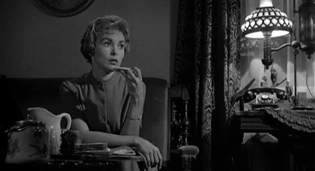 Psycho movie still: medium — A dimly lit room features a patterned curtain and a desk with a lamp. A woman in a blouse sits at a table and eats a…