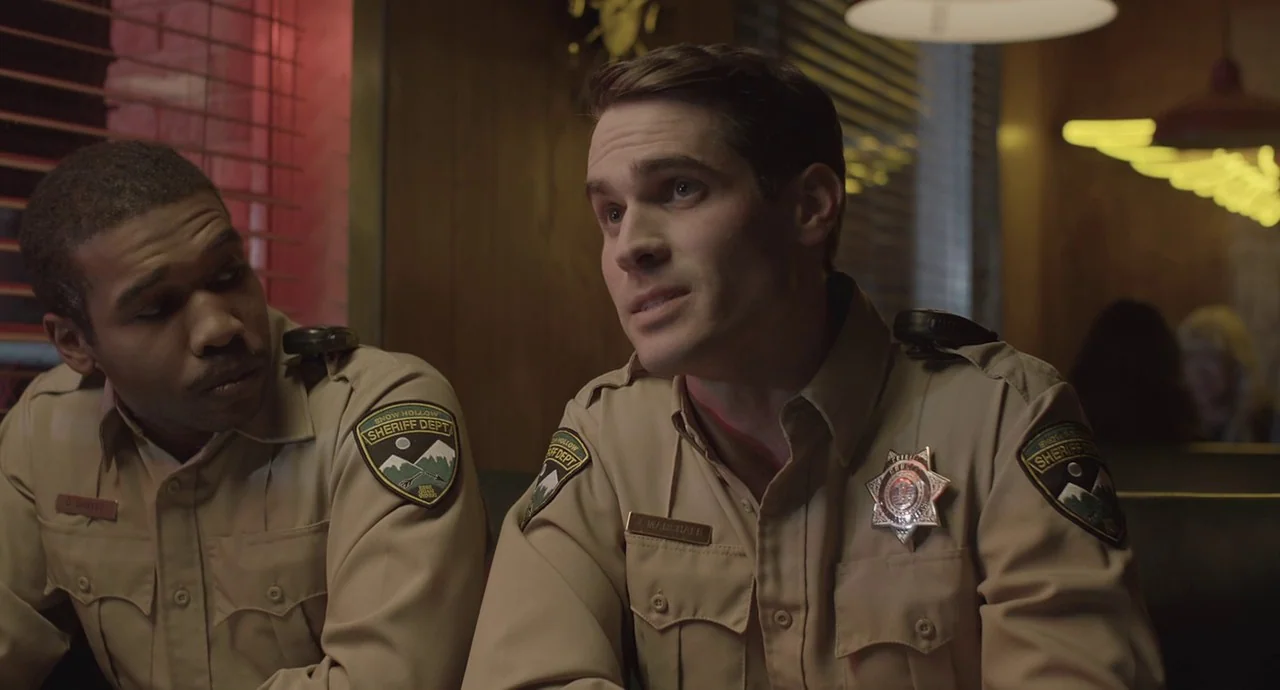 The Wolf of Snow Hollow (2020) movie still: medium — Two officers wearing uniform shirts sit inside a booth at a local diner. The men converse while…
