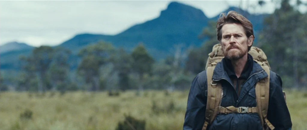 The Hunter (2011) movie still: medium — An open grassy field stretches toward distant mountains under an overcast sky. A man carrying a tan backpack…