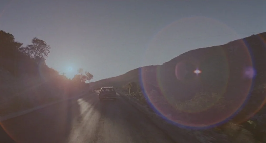 Y Tu Mamá También (2001) movie still: wide — An asphalt road winds through a rugged mountainous landscape during the late afternoon hours. A lone…