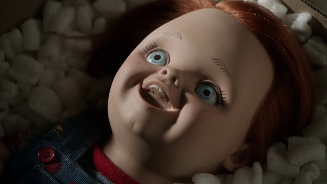 Curse of Chucky (2013) movie still: closeup — A plastic doll lies amidst white styrofoam packing peanuts inside a cardboard shipping box. The doll…