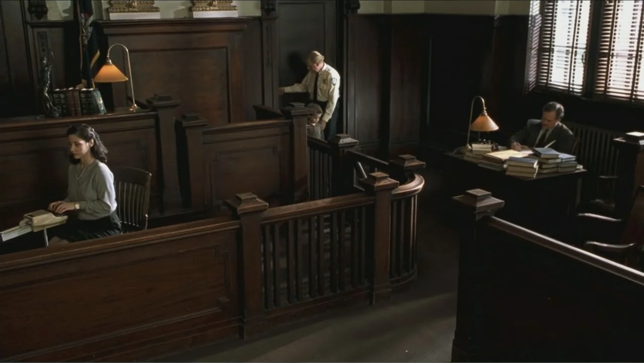 Big Daddy (1999) movie still: wide — A wood-paneled office inside a historic courtroom with heavy oak furniture and venetian blinds. A woman sits at…