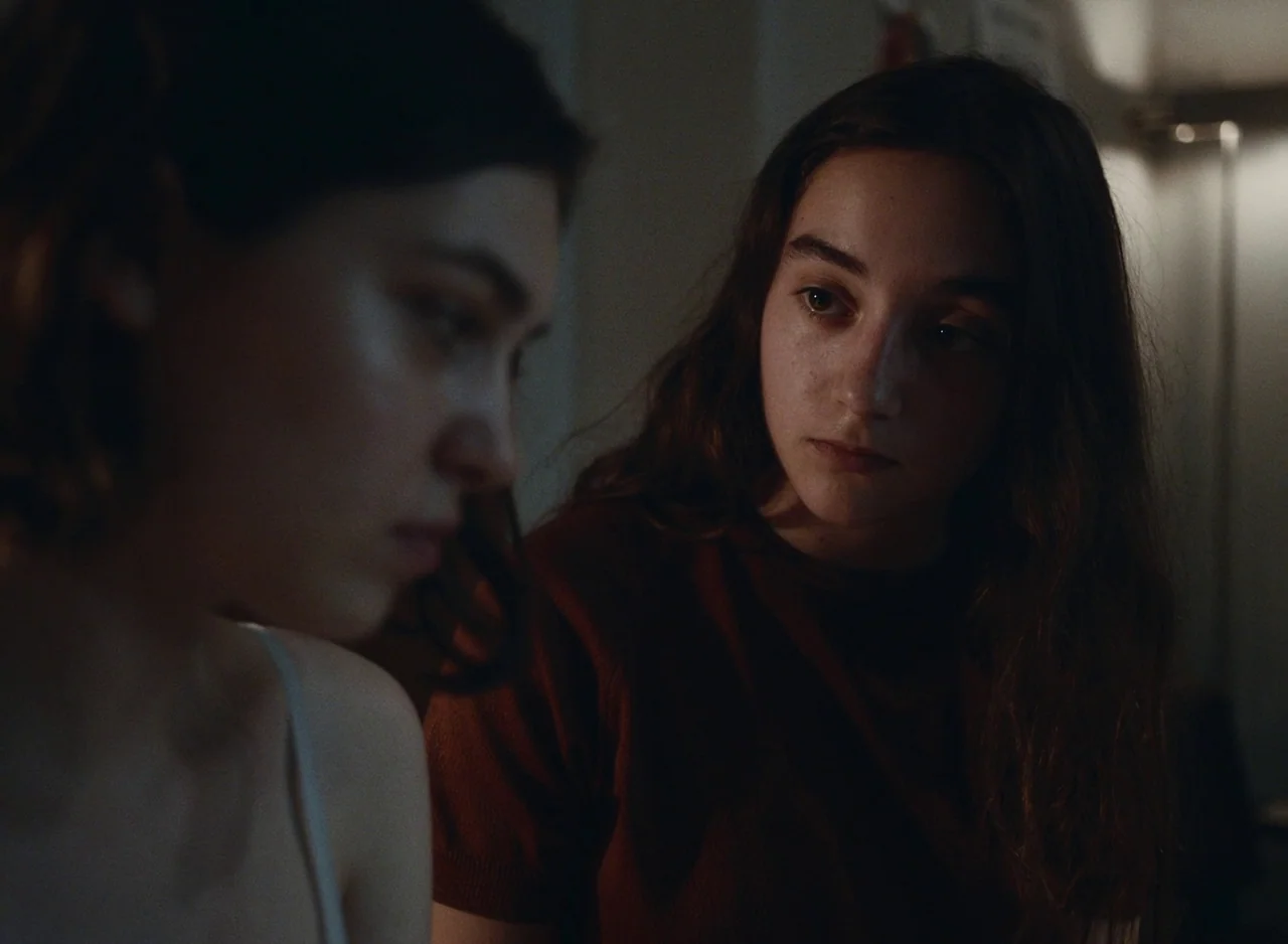 Happening (2021) movie still: medium — A dimly lit room with muted wall tones provides the backdrop for a quiet conversation. Two young women face…