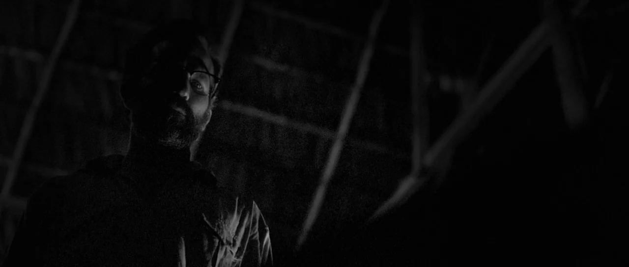 Embrace Of The Serpent (2015) movie still: medium — An interior space with wooden rafters visible in the dark background. A man stands below looking…