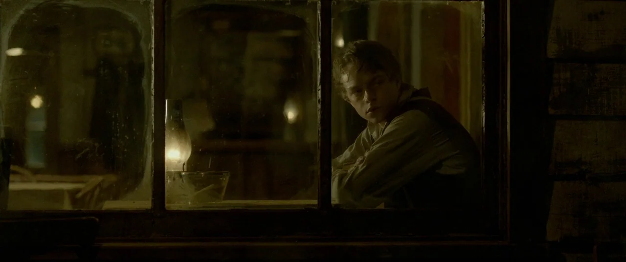 Lawless (2012) movie still: medium — A dimly lit room seen through a dark, multi-paned wooden window frame. A young man sits at a table near a…