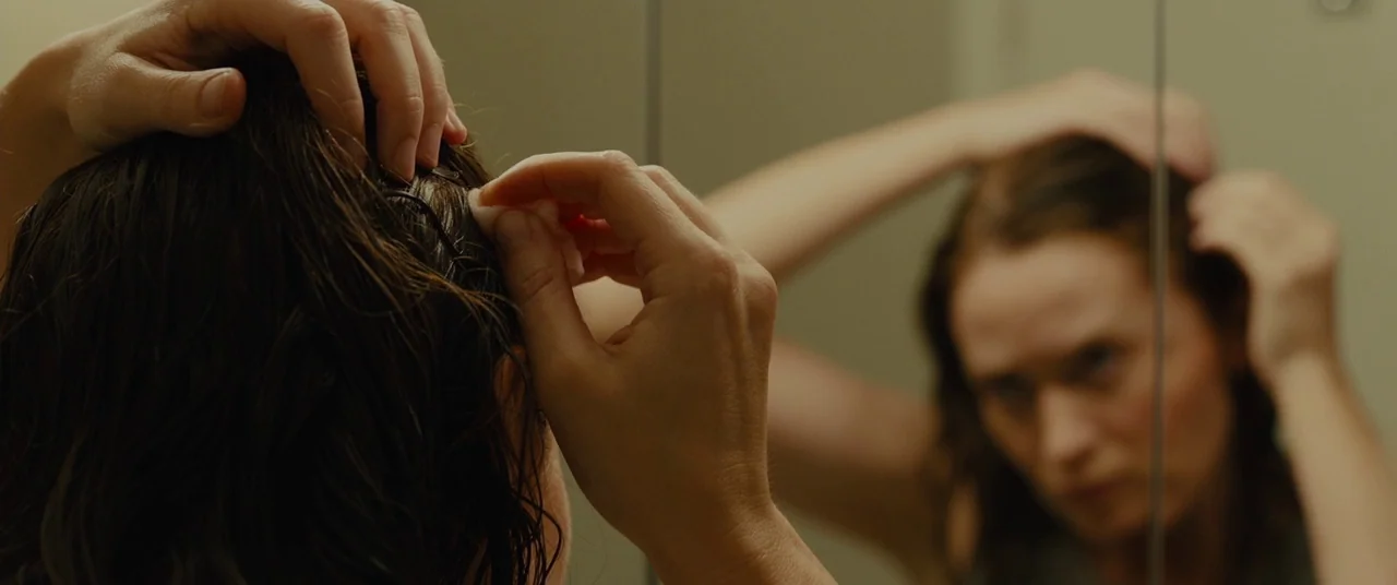 Sicario (2015) movie still: closeup — A woman stands before a glass mirror looking at her own reflection. She uses a small pad to attend to her wet…