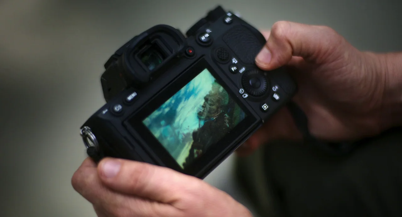 Civil War (2024) movie still: closeup — A pair of human hands hold a black mirrorless digital camera. The camera screen shows an image of a person…