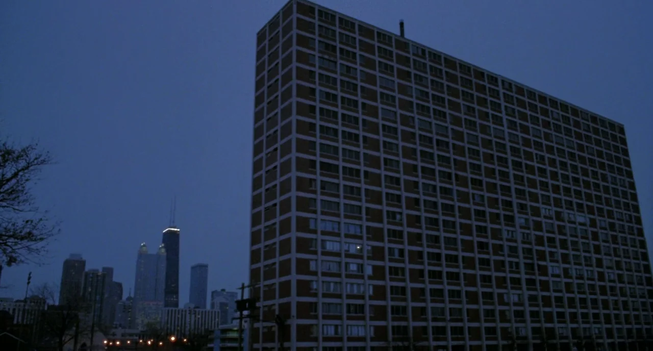 Candyman (1992) movie still: establishing — A tall, rectangular apartment block with a brick grid facade looms in the foreground. In the distance,…