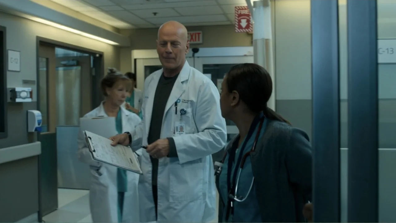 Death Wish (2018) movie still: medium — A sterile hospital corridor with room number signs and fluorescent lighting on the ceiling. A male doctor in…
