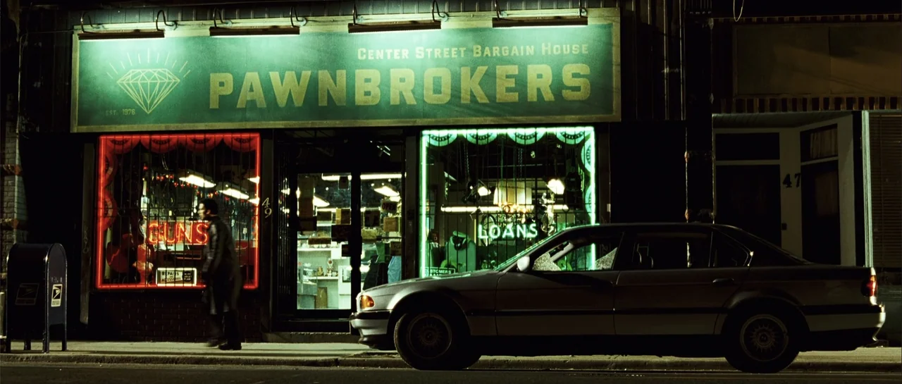 Shoot 'Em Up (2007) movie still: wide — A pawn shop at night features a large green diamond-patterned sign above the storefront windows. A man in a…
