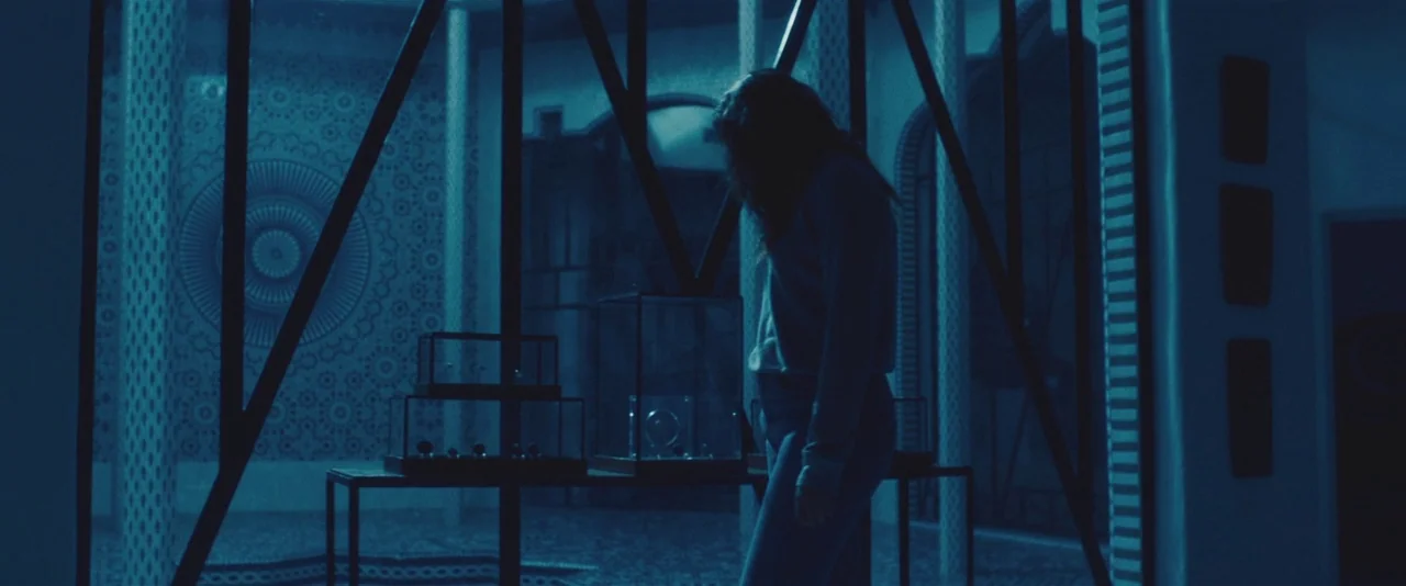 Gemini (2017) movie still: medium — A dark interior with patterned tile walls features a low-profile glass display exhibit. A young woman stands in…