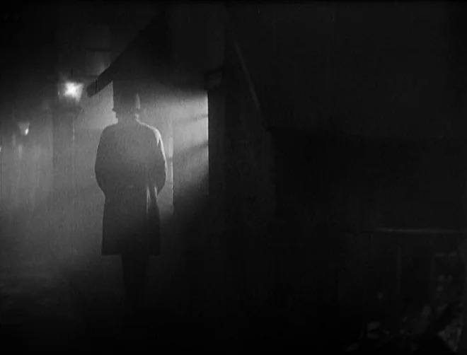 Pandora’s Box (1929) movie still: medium — A dark, foggy urban street at night with stone walls and dim streetlights. A man in a long coat and hat…