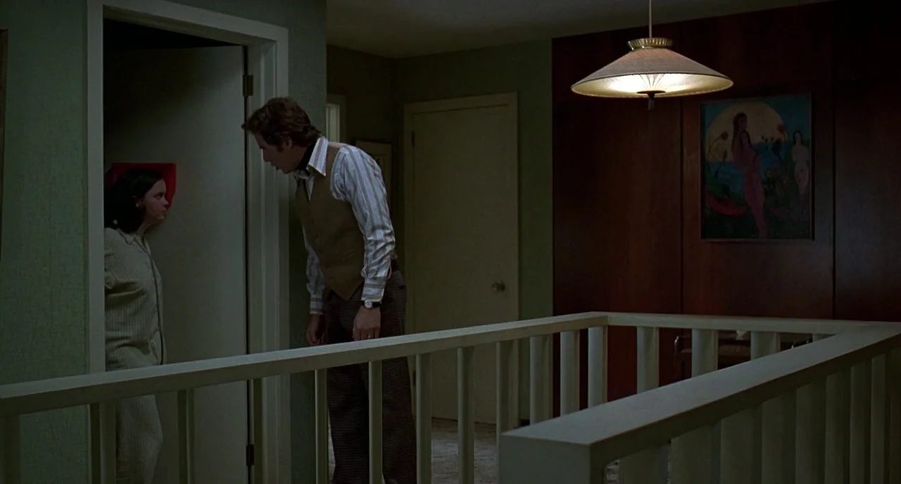 The Ice Storm (1997) movie still: medium — A residential landing with green textured walls and dark wood paneling at night. A man faces a woman who…