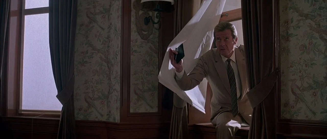 A View to a Kill (1985) movie still: medium — A classic room features patterned floral wallpaper and heavy brown curtains flanking a window. A man…