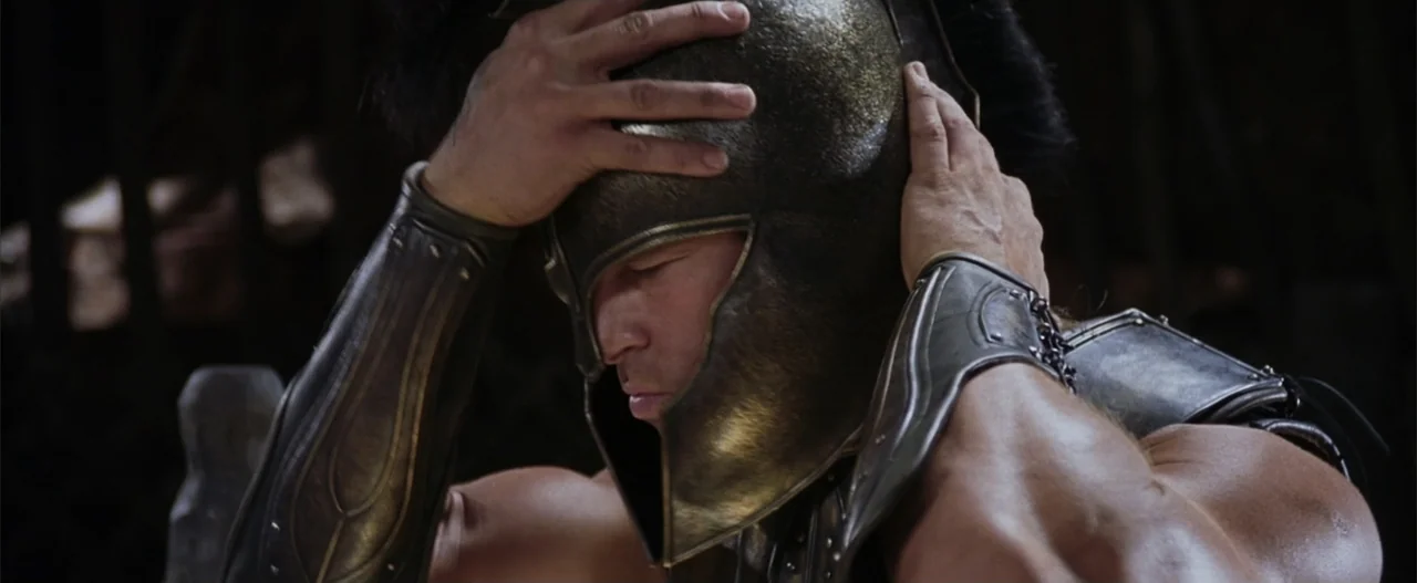 Troy (2004) movie still: closeup — A man stands outdoors wearing an ancient bronze helmet and forearm armor plates. He holds his hands against the…
