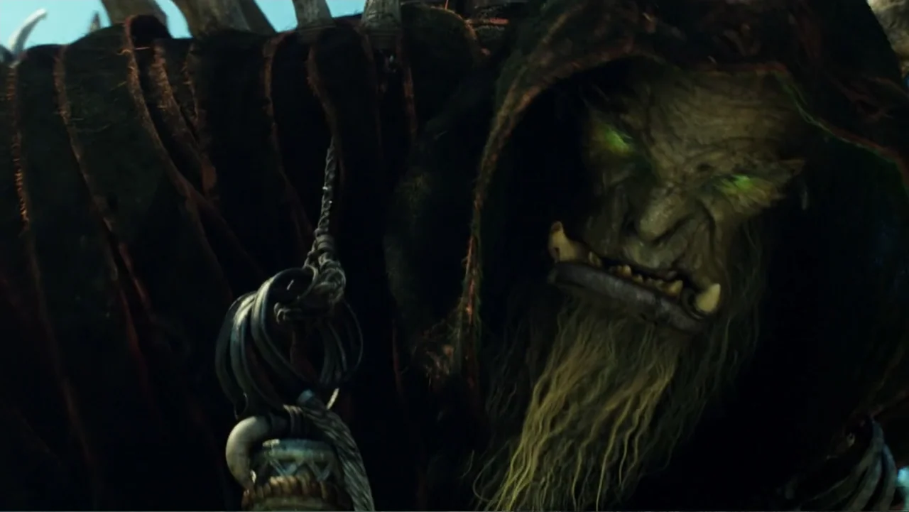Warcraft (2016) movie still: closeup — A dark fantasy environment with an aged orc wearing a tattered dark hood. The orc stares forward while…