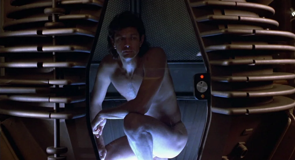 The Fly (1986) movie still: medium — A dark laboratory interior featuring a metallic pod with circular tiered ribs. A man crouches inside the…