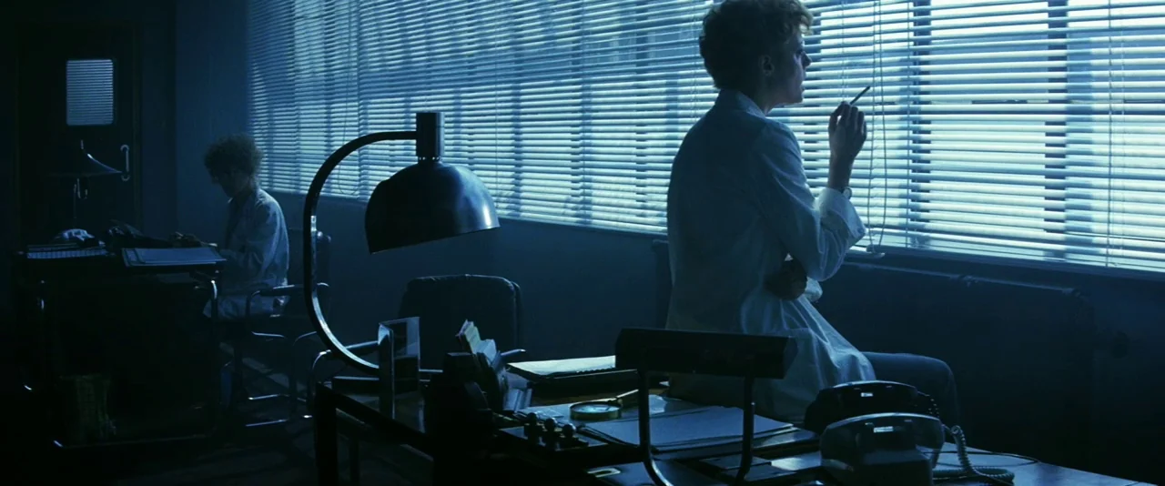The Hunger (1983) movie still: medium — A dimly lit room features rows of windows covered by horizontal blinds. Two individuals in white coats…