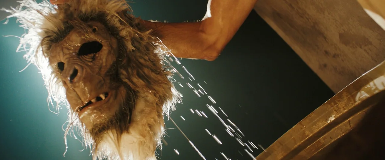 Monkey Man (2024) movie still: closeup — A wet primate mask with coarse synthetic fur is being rinsed under shower water. A person's arm holds the…