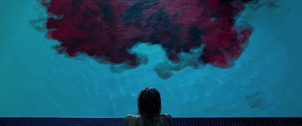 It Follows (2015) movie still: medium — A lone person rests at the edge of a pool filled with blue water. A large cloud of deep red ink disperses…